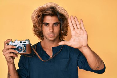 Şapkalı genç İspanyol bir adam elinde eski bir kamerayla elinde dur işareti ile ciddi ve kendinden emin bir ifadeyle savunma jesti yapıyor. 