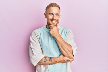 Elleri çenesinde gündelik kıyafetler giyen genç beyaz bir adam soru soruyor, düşünceli bir yüz ifadesi var. Düşünceli bir yüzle gülümsüyor. şüphe kavramı. 