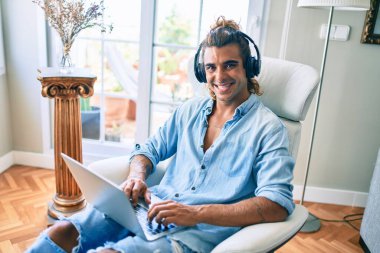 Evde dizüstü bilgisayar ve kulaklık kullanarak mutlu gülümseyen genç İspanyol adam.