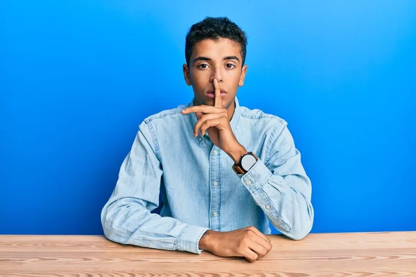 若いハンサムなアフリカ系アメリカ人の男性は テーブルの上に座ってカジュアルな服を着て唇に指で静かになるように求めている 沈黙と秘密の概念 — ストック写真