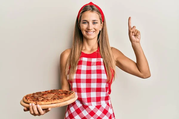 Młoda Blondynka Nosząca Profesjonalny Fartuch Piekarski Trzymająca Włoską Pizzę Uśmiechnięta — Zdjęcie stockowe