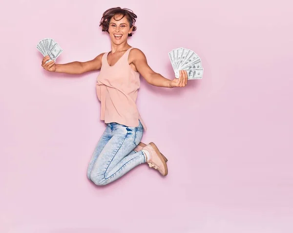 若い美しい女性は幸せな笑みを浮かべてドル紙幣を保持 孤立したピンクの背景の上に顔に笑顔でジャンプ — ストック写真