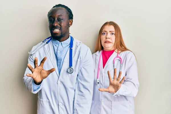 Young interracial couple wearing doctor uniform and stethoscope disgusted expression, displeased and fearful doing disgust face because aversion reaction. with hands raised