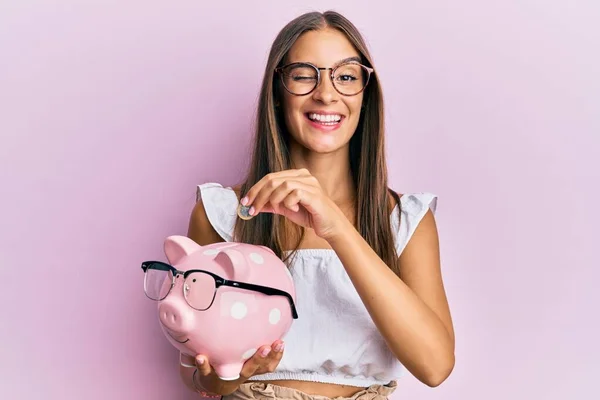 Junge Hispanische Frau Hält Sparschwein Mit Brille Und Münze Zwinkernd — Stockfoto