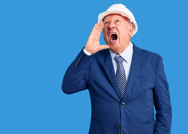 Senior handsome grey-haired man wearing suit and architect hardhat shouting and screaming loud to side with hand on mouth. communication concept.