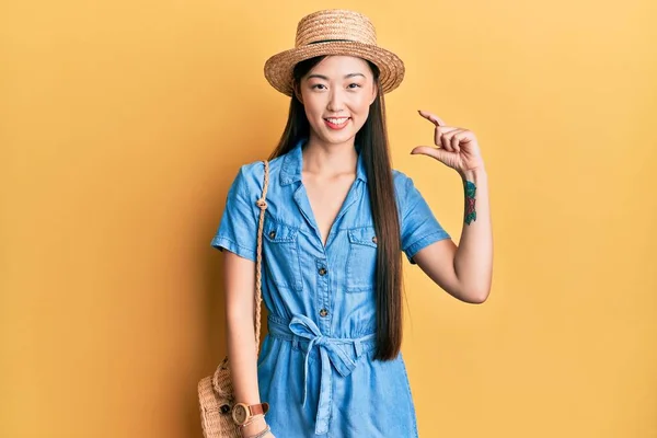 Young chinese woman wearing summer hat smiling and confident gesturing with hand doing small size sign with fingers looking and the camera. measure concept.