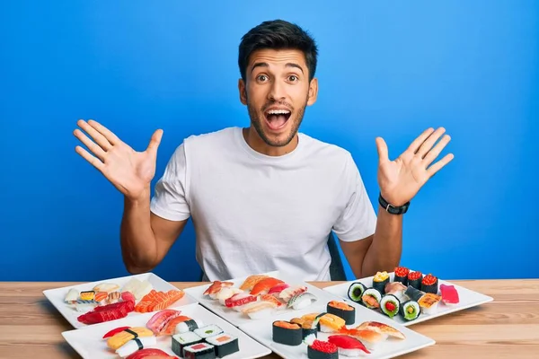 Young handsome man eating sushi sitting on the table celebrating crazy and amazed for success with arms raised and open eyes screaming excited. winner concept