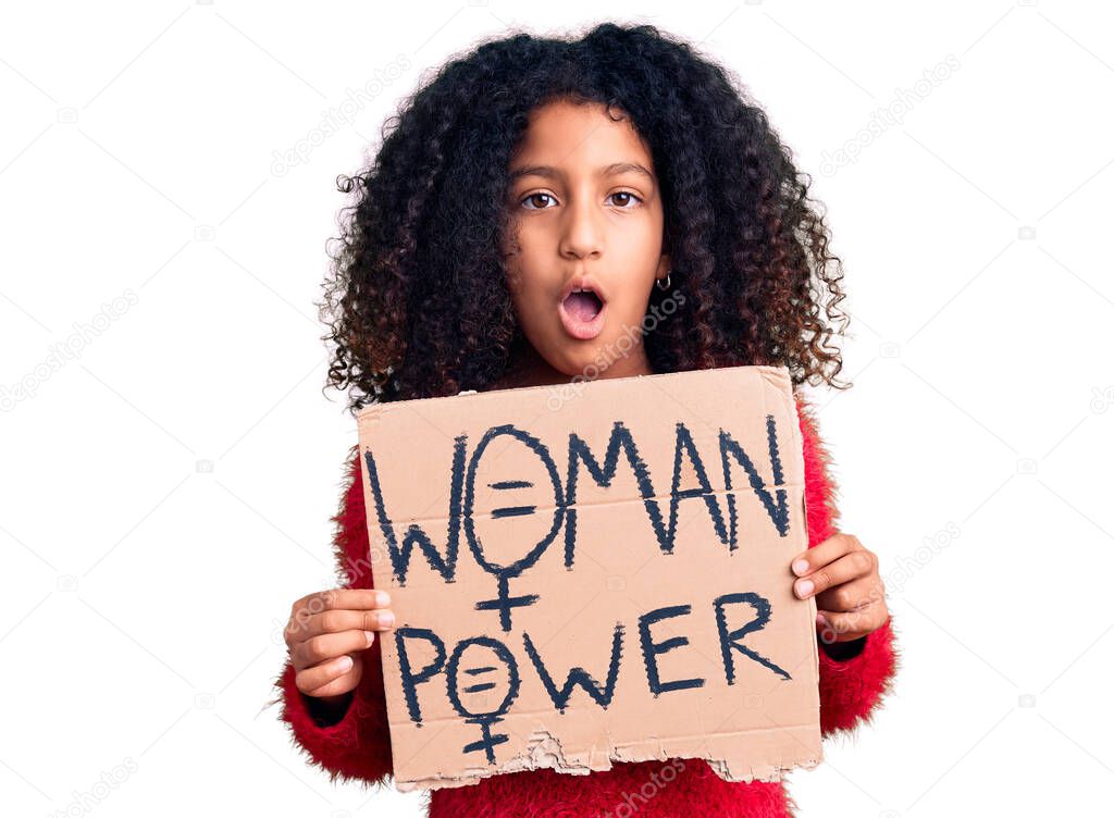 African american child with curly hair holding woman power banner scared and amazed with open mouth for surprise, disbelief face 