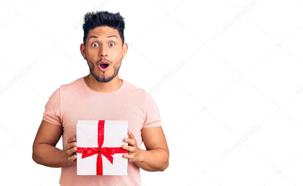 Handsome latin american young man holding gift scared and amazed with open mouth for surprise, disbelief face 