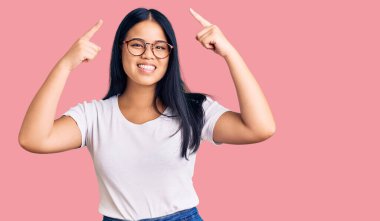 Genç, güzel Asyalı bir kız, günlük kıyafetler ve gözlük takıyor, gülümsüyor iki eliyle başını işaret ediyor, harika bir fikir ya da düşünce, iyi bir hafıza. 