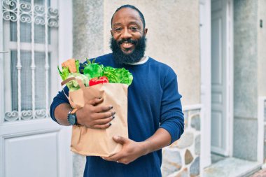 Sakallı Afrikalı Amerikalı bir adam elinde süpermarketten alınmış kağıt poşetler tutuyor.