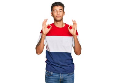 Genç, yakışıklı, Afro-Amerikalı, günlük kıyafetler giymiş rahat ve gözleri kapalı gülümseyen parmaklarıyla meditasyon hareketi yapan bir adam. yoga konsepti. 