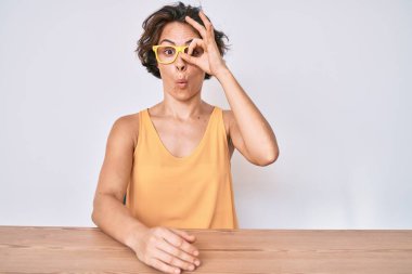 Gündelik giysiler ve gözlük takan genç İspanyol kadın masaya oturmuş şaşırmış yüz ifadesiyle şaşırmış bir şekilde parmaklarının arasından bakıyor. İnançsız ifade. 