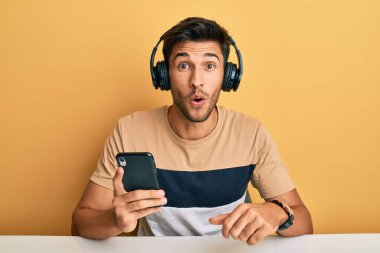 Akıllı telefon kullanan, kulaklık takan, ağzı açık, korkmuş ve şaşırmış genç bir adam. 