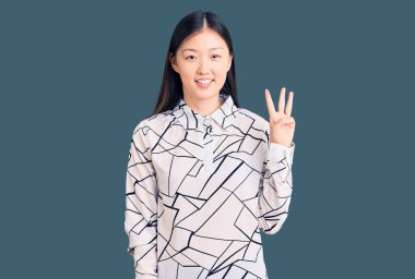 Genç, güzel, Çinli bir kadın, rahat bir tişört giyip üç numaralı parmaklarıyla işaret ederken, kendinden emin ve mutlu bir şekilde gülümsüyor.. 