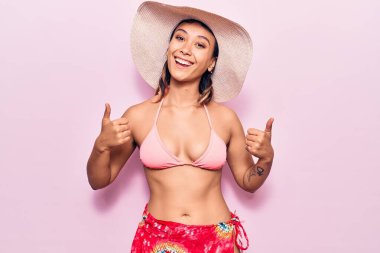 Bikini giyen ve şapka takan genç bir kadın elleriyle olumlu jest yapıyor, gülümsüyor ve mutlu. Neşeli ifade ve kazanan jest. 