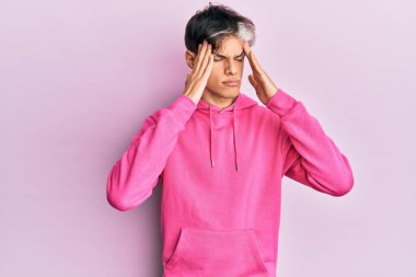 Latin asıllı genç bir adam, günlük pembe kazak giyiyor, elleri kafasında, stresten baş ağrısı çekiyor. migren ağrısı. 