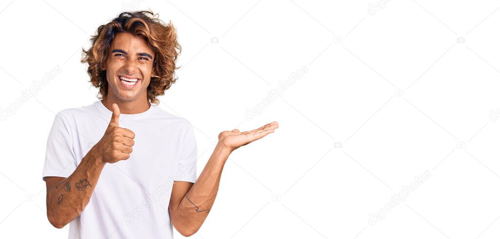 Young hispanic man wearing casual white tshirt showing palm hand and doing ok gesture with thumbs up, smiling happy and cheerful 