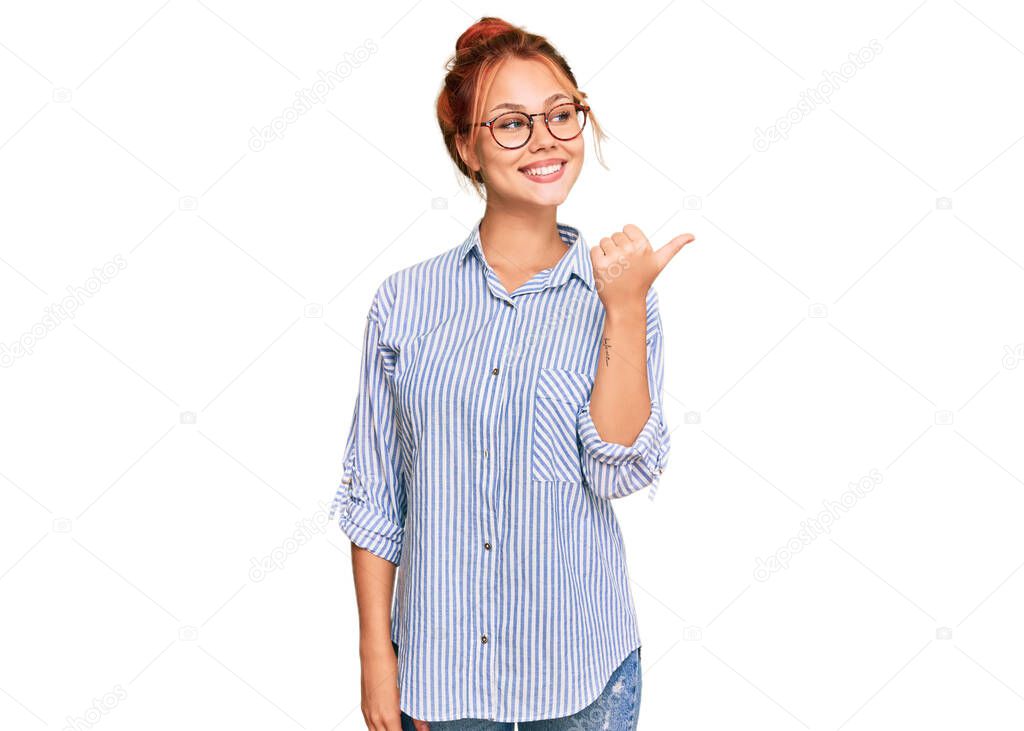 Young redhead woman wearing casual clothes and glasses smiling with happy face looking and pointing to the side with thumb up. 