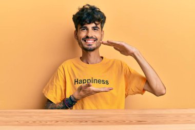 Üzerinde mutluluk mesajı olan tişört giyen genç İspanyol adam masaya oturmuş büyük ve büyük boyut işareti, ölçü sembolü gösteren el hareketleriyle işaret ediyor. Kameraya bakıp gülümsüyor. ölçüm kavramı. 