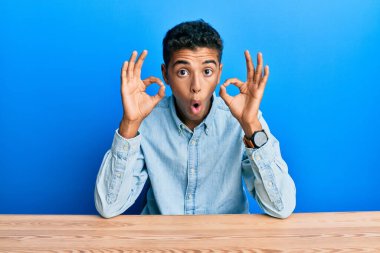 Genç, yakışıklı, Afro-Amerikalı, günlük giysiler içinde masada oturmuş, şaşırmış ve şok olmuş bir şekilde parmaklarıyla onay sembolü çiziyor. çılgın ifade 