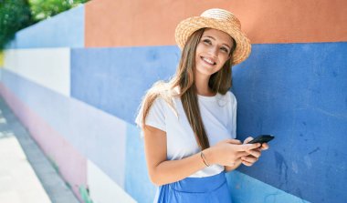 Tatildeki genç sarışın kadın mutlu bir şekilde gülümsüyor. Akıllı telefon kullanıyor ve şehrin sokaklarında duvara yaslanıyor.