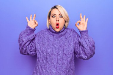 Güzel sarışın ve boyunda bir kadın mor arka plan üzerine balıkçı yaka kazak giyiyor. Şaşırmış ve şaşırmış görünüyor. Tamam sembolünü parmaklarıyla yapıyor. Çılgın ifade