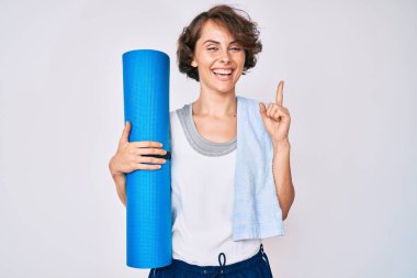 Elinde yoga minderi tutan genç İspanyol kadın bir fikir veya soruyla gülümsüyor ve mutlu bir yüzle işaret ediyor. 