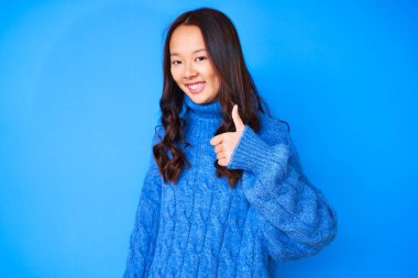 Kış süveteri giyen genç, güzel bir Çinli kız mutlu bir el hareketi yapıyor. Kameraya bakıp başarıyı gösteren ifade. 