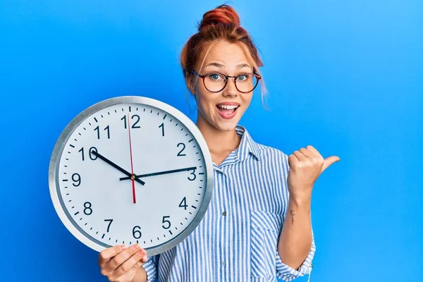 Joven Pelirroja Sosteniendo Gran Reloj Apuntando Pulgar Hacia Lado Sonriendo —  Fotos de Stock