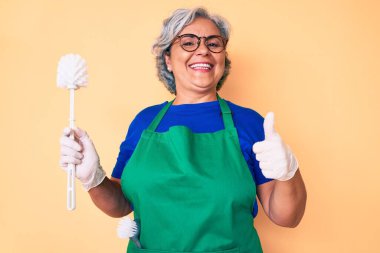 Önlük takan kıdemli İspanyol kadın elinde fırça, mutlu ve olumlu gülümsüyor, başparmağıyla mükemmel ve onay işareti yapıyor. 