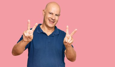 Orta yaşlı, kel, sıradan kıyafetler giyen, gülümseyen ve parmaklarını zafer işareti yaparken gösteren bir adam. İki numara.. 