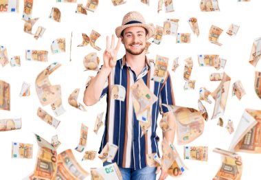 Yazlık şapka takan yakışıklı beyaz adam üç numarayı gösterip işaret ederken kendinden emin ve mutlu bir şekilde gülümsüyor..