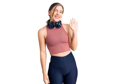 Güzel beyaz kadın spor kıyafetleri giyip kulaklık takıyor ve merhaba diyor. Mutlu ve güler yüzlü, dostça bir jest. 