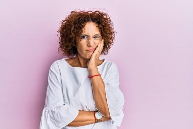Güzel, orta yaşlı olgun bir kadın pembe arka planda şık elbiseler giyiyor yorgun görünüyor ve kollarını kavuşturarak depresyondan sıkılmış görünüyor.. 
