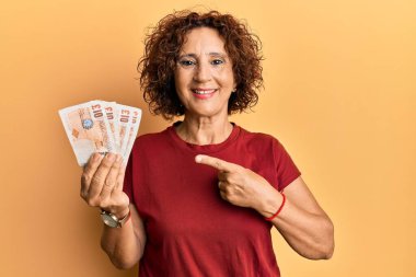 Güzel, orta yaşlı, olgun bir kadın elinde 10 pound banknotla mutlu bir şekilde el ve parmak işareti yapıyor. 