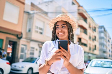 Güneşli bir yaz gününde açık havada akıllı telefon kullanarak mutlu bir şekilde gülümseyen Afrikalı Amerikalı genç bir kadın.