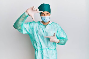 Cerrah üniforması ve tıbbi maske takan genç yakışıklı adam gülümseyerek elleri ve parmakları gülen yüzüyle çerçeve yapıyor. yaratıcılık ve fotoğrafçılık kavramı. 