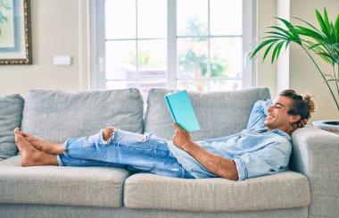 Genç İspanyol gülümseyen mutlu bir kitap okuyucu evdeki kanepede uzanıyor.