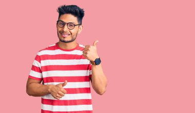 Yakışıklı Latin Amerikalı gündelik giysiler ve gözlük takmış arka tarafı işaret eden el ve başparmakları havada, kendinden emin gülümseyen genç adam. 