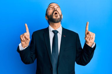 Uzun saçlı, sakallı, takım elbiseli ve kravatlı çekici bir adam şaşırmış ve şaşırmış bir şekilde yukarı bakıyor ve parmaklarıyla işaret ediyor ve kollarını kaldırıyor.. 