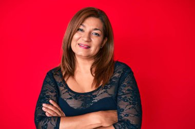 Orta yaşlı esmer, zarif elbiseler giyen, kırmızı arka planda mutlu yüzlü, kollarını kavuşturup kameraya bakan bir kadın. pozitif kişi. 