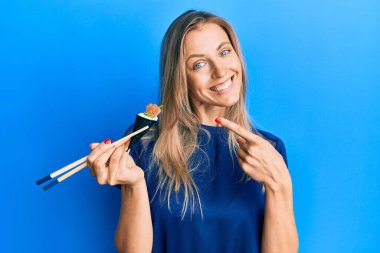 Güzel beyaz kadın temaki suşisi yiyor yemek çubuklarıyla gülümsüyor mutlu bir şekilde eli ve parmağı işaret ediyor. 