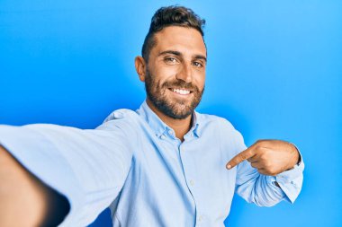 Sakallı yakışıklı bir adam akıllı telefonuyla fotoğraf çekiyor. Kendini mutlu ve gururlu bir şekilde gösteren birini işaret ediyor. 