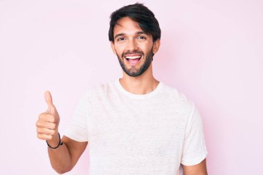 Yakışıklı, beyaz tişört giyen mutlu ve pozitif gülümseyen, başparmağıyla mükemmel ve onay işareti yapan bir İspanyol. 
