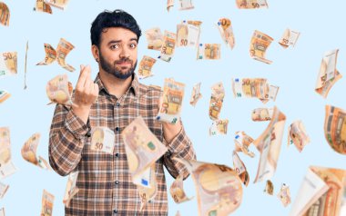 Gündelik kıyafetler giyen genç İspanyol adam elleriyle para hareketi yapıyor, maaş istiyor, milyonerlik işi yapıyor.