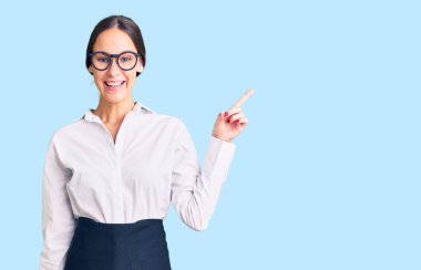 Güzel, esmer, profesyonel garson önlüğü giyen, yüzünde kocaman bir gülümseme olan genç bir kadın. Elinle kameraya bakıyor.. 