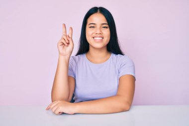 Genç, güzel Asyalı bir kız, günlük giysiler içinde masaya oturmuş, gülümsüyor ve kameraya göz kırpıyor zafer işareti yapıyor. İki numara.. 