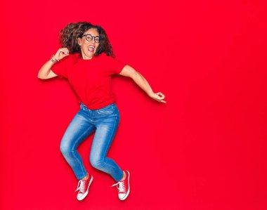 Orta yaşlı, güzel İspanyol bir kadın günlük kıyafetler ve mutlu bir gülümseme takınıyor. İzole edilmiş kırmızı arka planda yüzünde gülümsemeyle zıplıyor