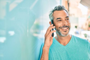 Orta yaşlı, kır saçlı, akıllı telefondan konuşan ve şehrin duvarına yaslanan bir adam..